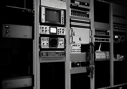 A collection of vintage electronic equipment is mounted in tall racks, featuring a variety of dials, knobs, and screens. Each unit appears to be a specialized piece of technology, possibly related to broadcasting or audio engineering. Cables are visibly hanging down the side of one rack.