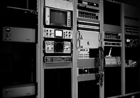 A collection of vintage electronic equipment is mounted in tall racks, featuring a variety of dials, knobs, and screens. Each unit appears to be a specialized piece of technology, possibly related to broadcasting or audio engineering. Cables are visibly hanging down the side of one rack.