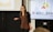 A woman with long hair and glasses is giving a presentation. She is holding a small remote or clicker in her right hand and standing in front of a projector screen and another screen displaying a logo with the word 'ACCELERATE'. She is wearing a black cardigan over a brown top.