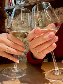 Two hands holding wine glasses in a toast, with brick wall background and shelves above. Both hands have rings, suggesting a celebratory occasion.