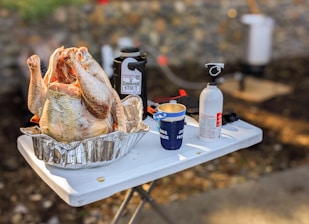 A raw turkey placed in a foil pan on a folding table outdoors. Nearby, there is a bottle labeled with a beer label, a mug with text, and a fire extinguisher. In the background, there is a blurry image of a fryer setup.