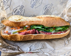 A freshly made sandwich with crispy baguette bread filled with green leafy vegetables, sliced red meat, and pickled vegetables, which includes carrots and onions. The sandwich is placed on branded wrapping paper.