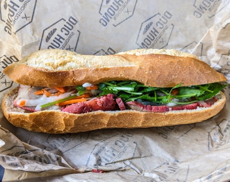 A freshly made sandwich with crispy baguette bread filled with green leafy vegetables, sliced red meat, and pickled vegetables, which includes carrots and onions. The sandwich is placed on branded wrapping paper.