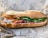 A freshly made sandwich with crispy baguette bread filled with green leafy vegetables, sliced red meat, and pickled vegetables, which includes carrots and onions. The sandwich is placed on branded wrapping paper.