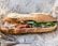 A freshly made sandwich with crispy baguette bread filled with green leafy vegetables, sliced red meat, and pickled vegetables, which includes carrots and onions. The sandwich is placed on branded wrapping paper.