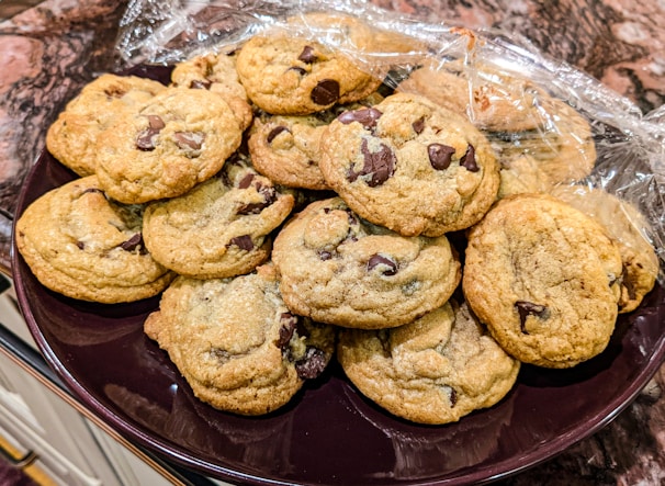 A plate piled with freshly baked chocolate chip cookies, wrapped partially with clear plastic wrap. The cookies are golden brown with visible chunks of chocolate, giving a warm and inviting appearance. The background features a marbled countertop, adding a touch of elegance to the presentation.