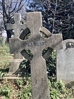 A weathered stone cross with the words 'AT REST' engraved on it stands prominently in a cemetery. The background features another cross and a traditional headstone surrounded by overgrown greenery and trees with bare branches.