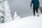 A person wearing winter clothing is standing on a snow-covered hill, holding a snowboard. Snow-laden trees are visible in the background, and the sky is overcast, suggesting a cold, wintery setting.