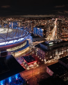 A nighttime cityscape featuring a large, illuminated stadium with a bright, domed roof. Surrounding the stadium are several modern buildings with bright lights, and a long stretch of highway with moving vehicles. The city is vibrant with numerous lights dotting the skyline.