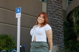 A woman with auburn hair smiles at the camera while standing outdoors. She is wearing a white top and green pants and is positioned next to a sign indicating wheelchair van accessibility. The background features a large palm tree and lush green plants, with the exterior of a building visible.