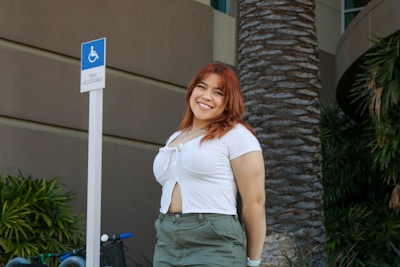 A woman with auburn hair smiles at the camera while standing outdoors. She is wearing a white top and green pants and is positioned next to a sign indicating wheelchair van accessibility. The background features a large palm tree and lush green plants, with the exterior of a building visible.
