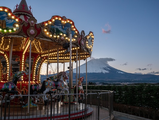 A whimsical carousel with ornate horses is brightly lit with string lights in the foreground. In the background, the iconic snow-capped Mount Fuji stands majestically under a clear blue sky, with a few scattered clouds.