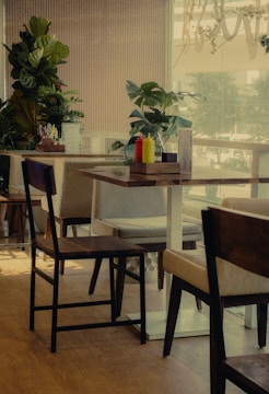A cozy café setting featuring wooden chairs and tables. The space is decorated with several green potted plants, creating a refreshing and natural ambiance. On the table, there are condiment bottles and a menu stand, with sunlight filtering through large windows providing a warm glow.
