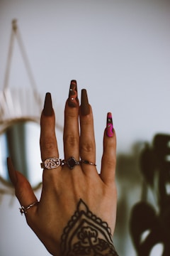 A hand with elaborately decorated nails featuring designs in brown and pink, adorned with small rhinestones. Several rings with intricate designs are worn on different fingers, and a detailed tattoo adorns the back of the hand. The background is blurred, emphasizing the hand and its embellishments.