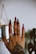 A hand with elaborately decorated nails featuring designs in brown and pink, adorned with small rhinestones. Several rings with intricate designs are worn on different fingers, and a detailed tattoo adorns the back of the hand. The background is blurred, emphasizing the hand and its embellishments.