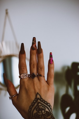 A hand with elaborately decorated nails featuring designs in brown and pink, adorned with small rhinestones. Several rings with intricate designs are worn on different fingers, and a detailed tattoo adorns the back of the hand. The background is blurred, emphasizing the hand and its embellishments.