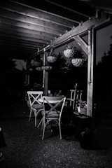 An outdoor patio setting featuring a wooden pergola with hanging flower baskets. Beneath the pergola, there are two wooden chairs and a table. The surrounding area is dimly lit, casting shadows and creating a serene evening ambiance.
