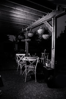 An outdoor patio setting featuring a wooden pergola with hanging flower baskets. Beneath the pergola, there are two wooden chairs and a table. The surrounding area is dimly lit, casting shadows and creating a serene evening ambiance.
