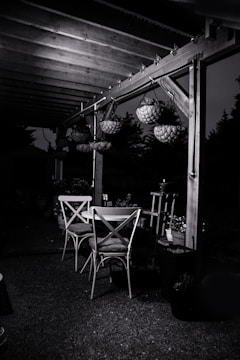 An outdoor patio setting featuring a wooden pergola with hanging flower baskets. Beneath the pergola, there are two wooden chairs and a table. The surrounding area is dimly lit, casting shadows and creating a serene evening ambiance.