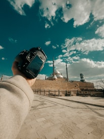 A person holding a DSLR camera in front of a grand architectural structure with multiple minarets and a dome under a partly cloudy sky. The foreground features a paved open area with light stone tiles.