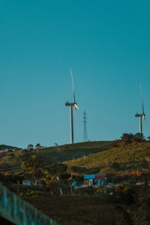 Two wind turbines standing on a green hillside under a clear blue sky. A small house with a blue roof is visible in the foreground surrounded by vegetation and trees. There is a power line or telephone pole near the wind turbines.