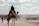 A person is riding a camel adorned with colorful blankets and decorations, overlooking a vast desert landscape. Several other people are walking nearby, with a horizon of distant city structures and a clear sky filled with clouds.