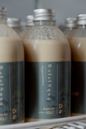 Several plastic bottles with metallic caps are filled with a creamy beige liquid. The bottles are labeled with a dark green-blue label reading 'BotolSqwy' and 'Kopisyu'. They are arranged closely together, hinting at a display or storage setting.