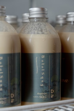 Several plastic bottles with metallic caps are filled with a creamy beige liquid. The bottles are labeled with a dark green-blue label reading 'BotolSqwy' and 'Kopisyu'. They are arranged closely together, hinting at a display or storage setting.
