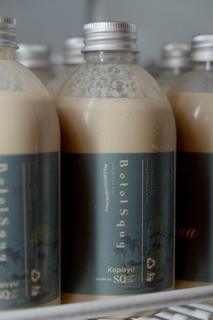 Several plastic bottles with metallic caps are filled with a creamy beige liquid. The bottles are labeled with a dark green-blue label reading 'BotolSqwy' and 'Kopisyu'. They are arranged closely together, hinting at a display or storage setting.