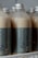 Several plastic bottles with metallic caps are filled with a creamy beige liquid. The bottles are labeled with a dark green-blue label reading 'BotolSqwy' and 'Kopisyu'. They are arranged closely together, hinting at a display or storage setting.