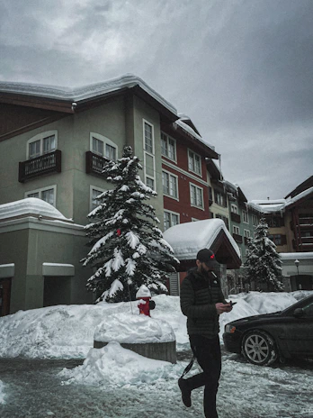 A snow-covered street scene features a man walking while looking at his phone. He is dressed warmly with a cap and jacket. Tall buildings with snow-laden roofs and windows surround the area, set against a cloudy gray sky. A snowy tree and a red fire hydrant are visible in the foreground, and a dark car is parked nearby.