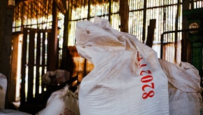 Large white sacks with red printed numbers are stacked in a dimly lit room. The background consists of wooden slats allowing light to filter through, casting a pattern of light and shadows. The environment suggests an industrial or storage setting.