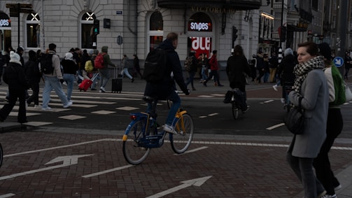A busy city intersection bustling with people crossing the street and a person riding a bicycle. The background features a large building with shops, including a clothing store and a sign that says 'Snipes'. People are dressed warmly, suggesting a cool climate, and some are carrying luggage.