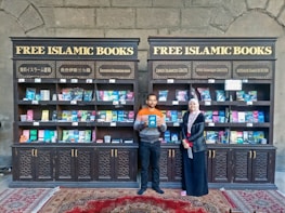 Two individuals are standing in front of two bookcases labeled 'Free Islamic Books.' The bookcases are filled with various books and pamphlets. The man is holding a book, and there is a carpet on the floor.