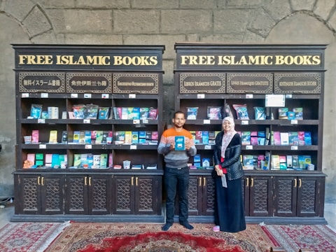 Two individuals are standing in front of two bookcases labeled 'Free Islamic Books.' The bookcases are filled with various books and pamphlets. The man is holding a book, and there is a carpet on the floor.