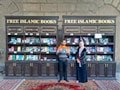 Two individuals are standing in front of two bookcases labeled 'Free Islamic Books.' The bookcases are filled with various books and pamphlets. The man is holding a book, and there is a carpet on the floor.