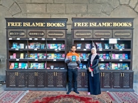 Two individuals are standing in front of two bookcases labeled 'Free Islamic Books.' The bookcases are filled with various books and pamphlets. The man is holding a book, and there is a carpet on the floor.