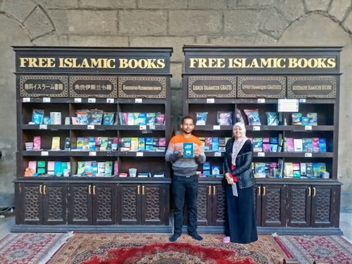 Two individuals are standing in front of two bookcases labeled 'Free Islamic Books.' The bookcases are filled with various books and pamphlets. The man is holding a book, and there is a carpet on the floor.