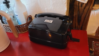 A vintage rotary telephone with a black glossy finish is placed on a red wooden surface. To the left is a wicker basket holding several bottles, including a clear one and a white one with blue labeling. A white electric kettle with a wooden handle is also visible. The background includes a wooden beam and a white textured wall.