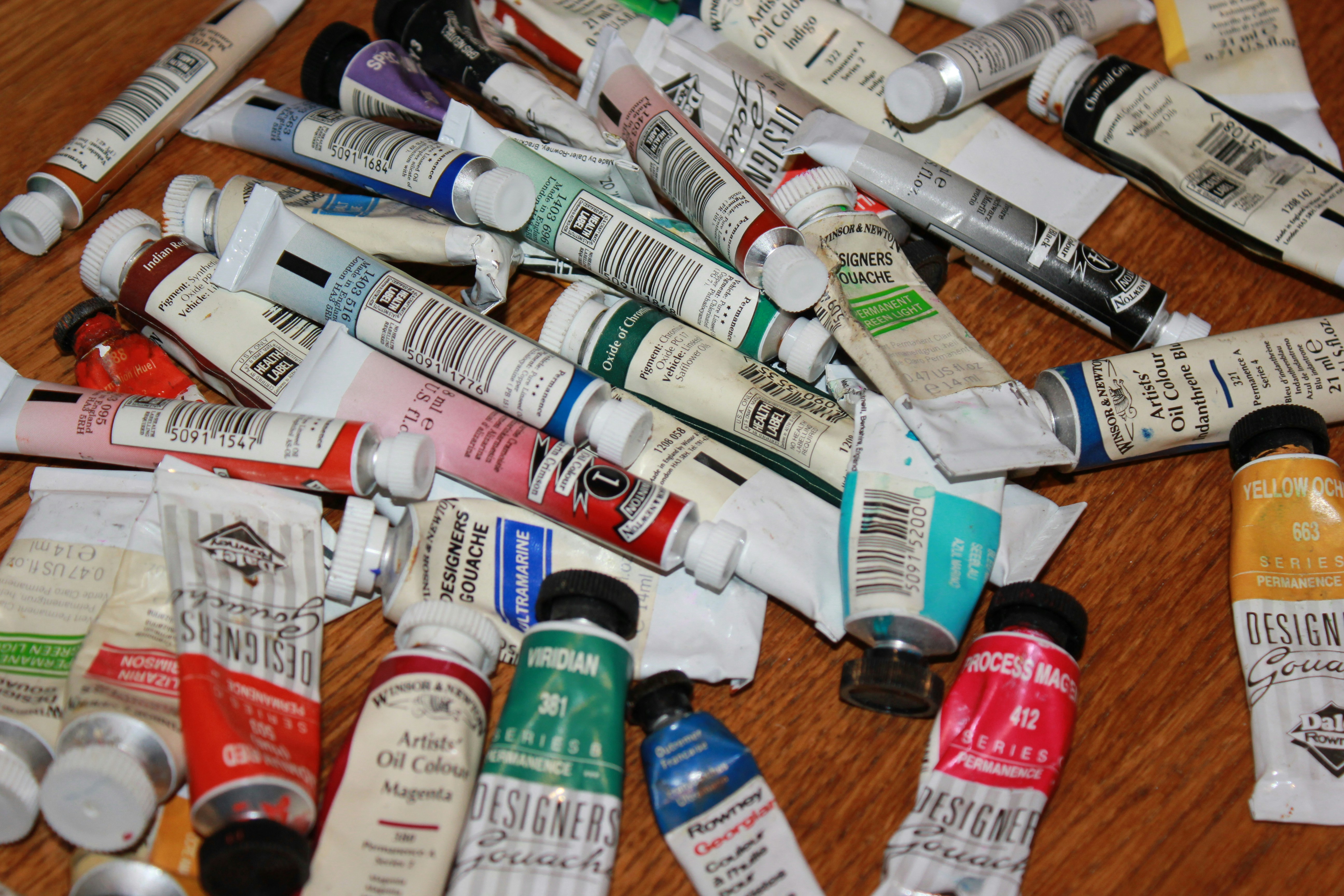 A collection of various paint tubes scattered across a wooden surface, featuring a mix of oil colors and gouache. The tubes are different sizes and brands, with labels indicating colors such as magenta, yellow ochre, and viridian.