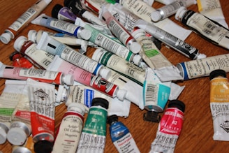 A collection of various paint tubes scattered across a wooden surface, featuring a mix of oil colors and gouache. The tubes are different sizes and brands, with labels indicating colors such as magenta, yellow ochre, and viridian.