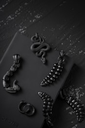 A collection of dark, intricately detailed jewelry pieces is arranged on a textured surface. The jewelry includes serpentine designs and other organic forms in a matte black finish.