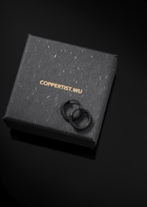 A pair of black textured rings is placed on top of a dark, sleek box. The box features the brand name 'COPPERTIST.WU' printed in gold letters.