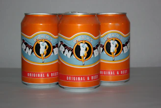 Three orange soda cans are placed in a row. The branding features a vintage illustration of a person in a white outfit, and the text on the label reads: 'Barr's' and 'Original & Best.' The background is plain and neutral, enhancing the focus on the cans.