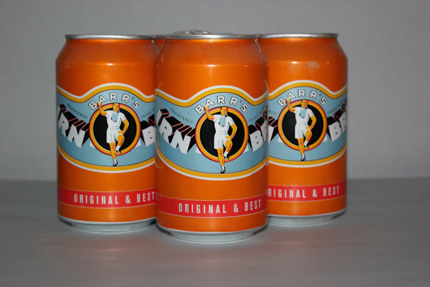 Three orange soda cans are placed in a row. The branding features a vintage illustration of a person in a white outfit, and the text on the label reads: 'Barr's' and 'Original & Best.' The background is plain and neutral, enhancing the focus on the cans.