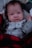 A baby is dressed in a light gray onesie with dark paw print patterns and a tag reading 'Daddy's Best Friend.' The baby is wrapped in a red and black buffalo plaid blanket, lying on a soft surface.