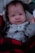 A baby is dressed in a light gray onesie with dark paw print patterns and a tag reading 'Daddy's Best Friend.' The baby is wrapped in a red and black buffalo plaid blanket, lying on a soft surface.