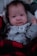 A baby is dressed in a light gray onesie with dark paw print patterns and a tag reading 'Daddy's Best Friend.' The baby is wrapped in a red and black buffalo plaid blanket, lying on a soft surface.