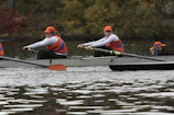 A team of rowers wearing orange caps and blue and orange uniforms are paddling in a rowing boat on a body of water. The rowers are focused and appear to be synchronizing their movements. The background features some greenery with autumnal colors suggesting a scenic outdoor setting.