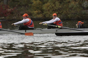 A team of rowers wearing orange caps and blue and orange uniforms are paddling in a rowing boat on a body of water. The rowers are focused and appear to be synchronizing their movements. The background features some greenery with autumnal colors suggesting a scenic outdoor setting.
